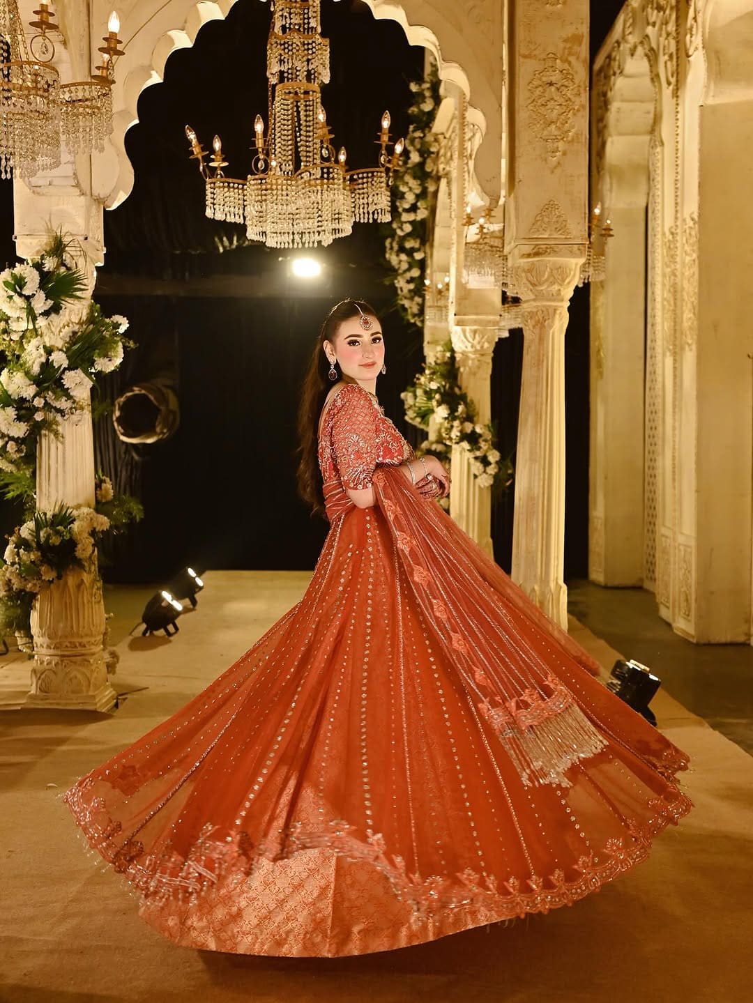 Coral pishwas in motion beneath a chandelier-lit arch